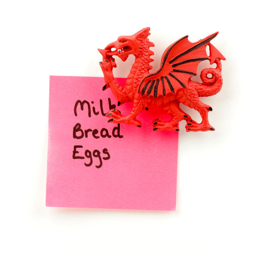 Angled overhead shot of a red Welsh Dragon magnet with black detailing, gripping a pink sticky note that displays a shopping list.