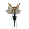 Detailed view of an ornate, bone-coloured skull bottle stopper, featuring prominent spikes and ridges. It has a metallic silver-coloured stem and black ribbed seal, inserted into a clear glass bottle neck.