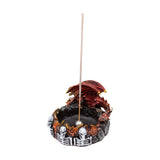 Angled view of a dark red, winged dragon incense burner, featuring skulls and orange flame details on its circular base, holding an unlit incense stick.