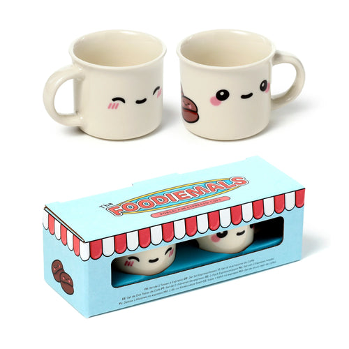 Angled view of two cream porcelain espresso cups with cartoon Foodiemals faces and handles, one showing closed eyes and the other an open-eyed coffee bean design. They are presented next to their light blue retail box, which features a red and white striped awning and 'Foodiemals Porcelain Espresso Cups' branding.