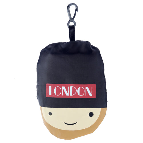 Front view of a handy foldable London souvenir shopping bag designed as a Guardsman's head, featuring a black top, a red band with white LONDON text, a beige face with eyes and a smile, and an attached black carabiner clip.