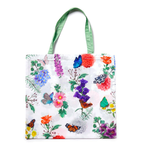 Front view of a white shopping tote bag featuring a vibrant multi-colored butterfly and meadow floral print, with green fabric handles, displayed on a white background.