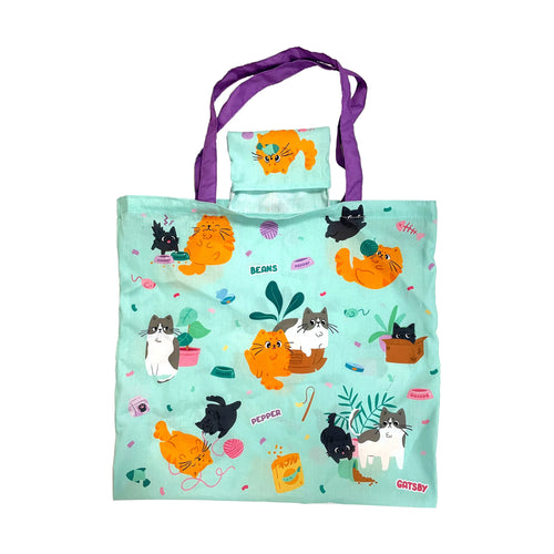 Front view of a foldable mint green shopping tote bag with purple handles, featuring a colourful cartoon pattern of various cats, cat toys, and food bowls with 'Beans', 'Pepper', and 'Gatsby' names.