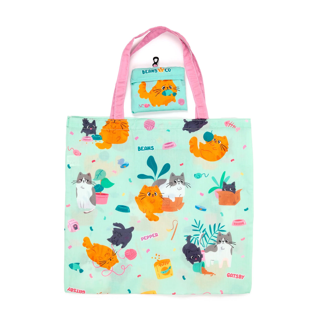 Front view of a light green foldable shopping tote bag with pink handles, featuring a playful colourful cartoon pattern of various cats and cat-themed accessories. A smaller 'Beans & Co' pouch with an orange cat design is clipped to the top.