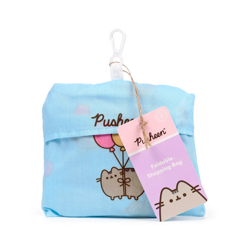 Front view of a light blue foldable shopping tote bag featuring Pusheen the Cat holding pink and yellow balloons. The compact bag has a white carabiner clip and a pink and purple product tag attached with twine.