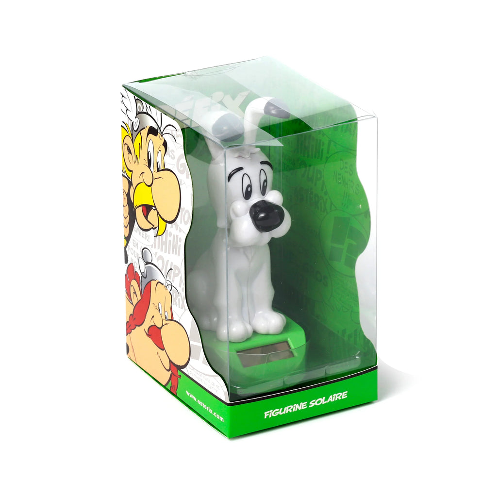 Front-side view of a collectable Asterix Idefix (Dogmatix) solar powered pal figurine. The white dog figure with black features is visible inside a clear plastic display box with green accents and character illustrations. It stands on a green base with a solar panel, on a white background.