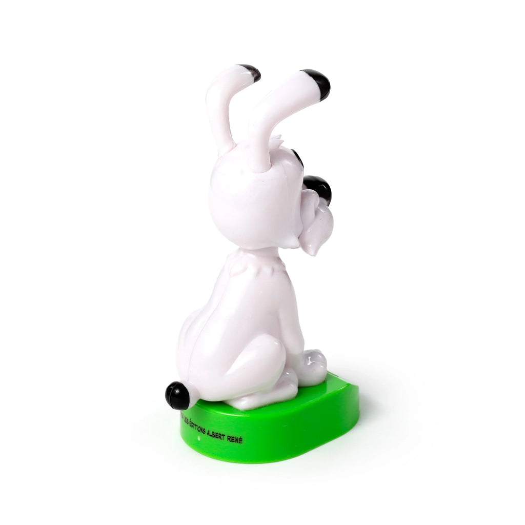Side-back view of a white Collectable Asterix Idefix solar pal, a dog figure with black-tipped ears and tail, seated on a green oval base.