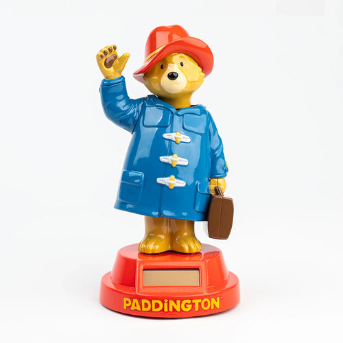 Front view of a Solar Pal Paddington Bear figurine, wearing a blue coat and red hat, waving and holding a brown briefcase. The red base features 'PADDINGTON' in yellow lettering and a solar panel.