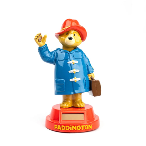 Solar Pal Paddington Bear figurine, front view, wearing a red hat and blue duffel coat with toggle fastenings. The yellow bear waves with one paw and holds a brown suitcase, standing on a red base with a visible solar panel and 'PADDINGTON' in yellow lettering.