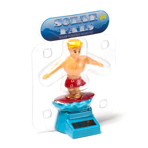 Front view of a Solar Pal figurine, depicting a blonde-haired male surfer character wearing red floral shorts, standing on a red surfboard. The surfboard is mounted on a blue wave-shaped base with a solar panel. The item is encased in clear plastic packaging with a blue header featuring SOLAR PALS and SOLAR POWERED NOVELTIES text.