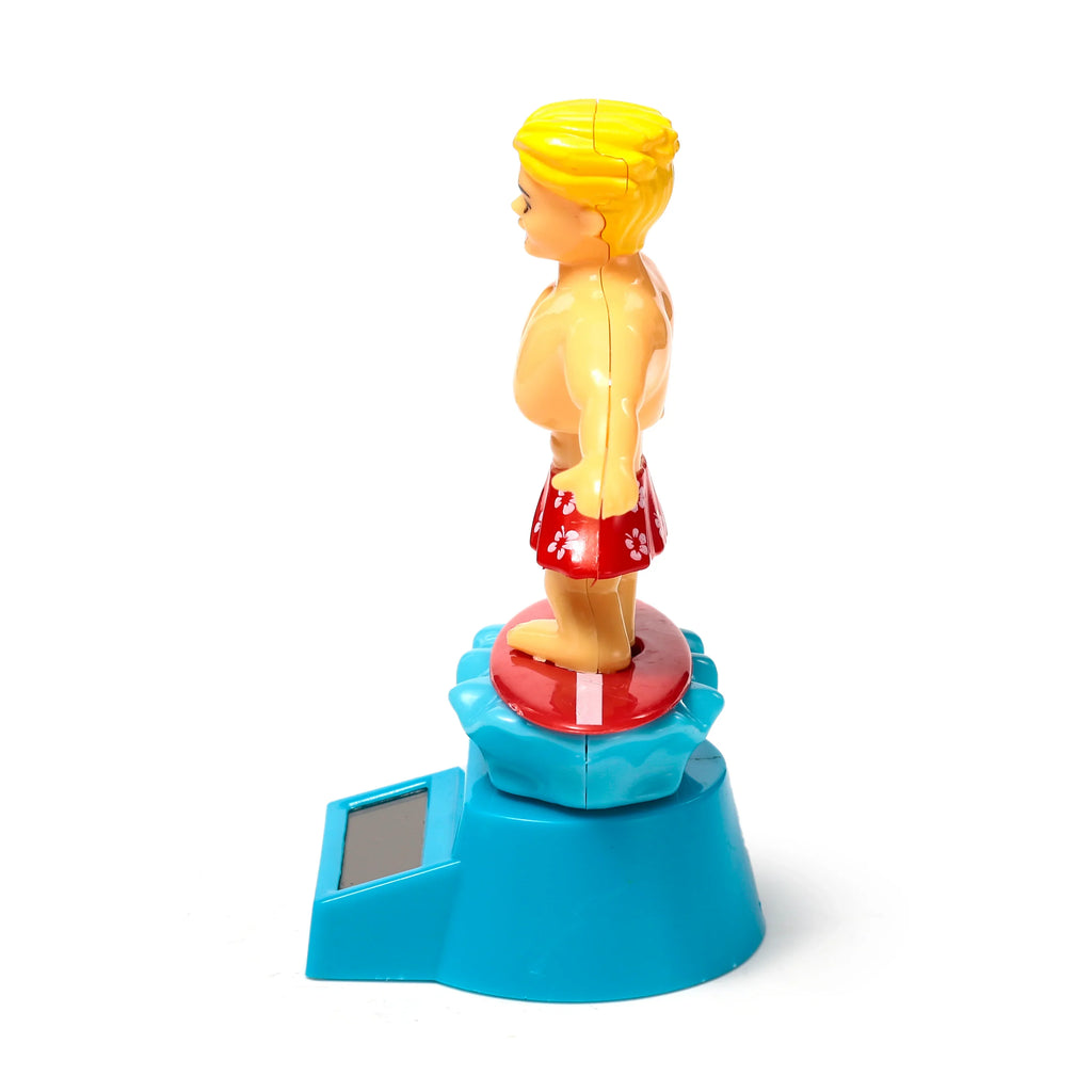 Side view of a Solar Pal Surger Dude figurine. The blonde-haired male wears red shorts with white floral patterns, standing on a red surfboard atop a blue base with a solar panel.