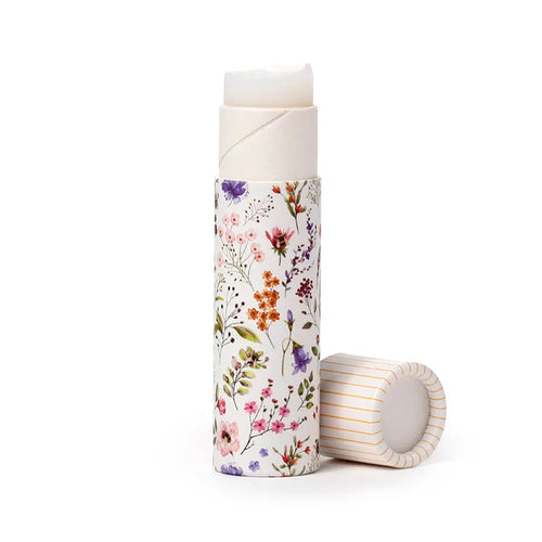 Front view of an open solid perfumed stick balm, a lip balm, in a white tube with a colorful floral pattern, and its striped cap lying beside it.