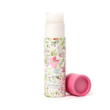 Front view of a solid perfumed stick balm in a white cylindrical tube with a pink botanical floral pattern. The light-colored balm stick is visible at the top, with a pink cap lying beside it.