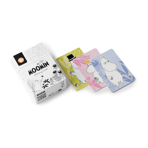 Angled view of a white memory card box with black Moomin character illustrations, 'Moomin Memory Cards' text, and an orange puzzle piece logo, beside three fanned-out character cards. The cards are yellow-green, pink, and light blue, each featuring a different Moomin character.