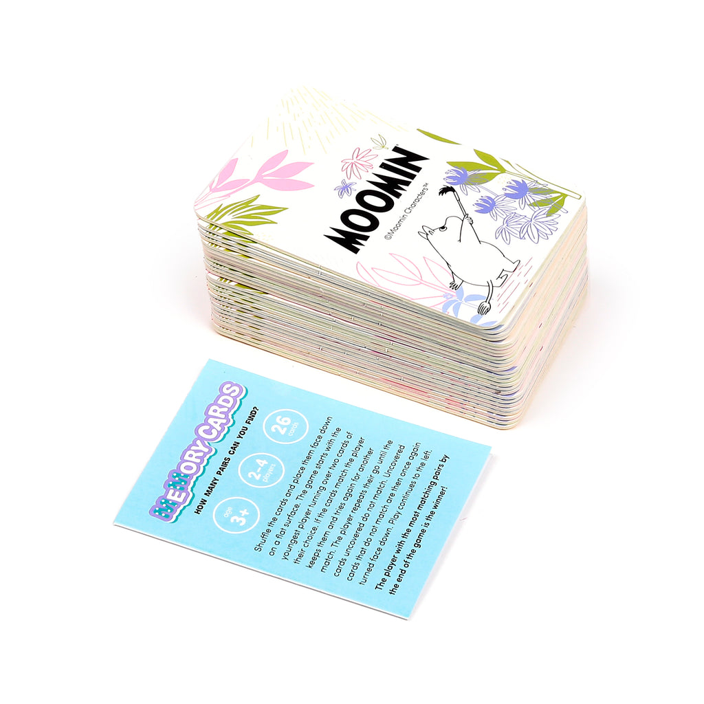 Angled view of a stack of children's memory cards, with the top card showing a Moomin character and pastel floral illustrations. Next to it is a light blue instruction card displaying 'Memory Cards' text, age 3+, 2-4 players, and 26 cards.