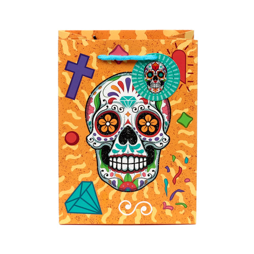 Front view of a medium Day of the Dead gift bag, featuring a vibrant orange background with a large, colourful sugar skull design. The skull has intricate floral patterns, flower eyes, and a diamond on its forehead. The bag is adorned with a purple cross, red heart, turquoise diamond, and scattered confetti shapes, complete with blue rope handles and a small circular tag showing another sugar skull.
