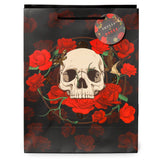 Front view of a black gift bag featuring a central white skull surrounded by vibrant red roses, with black rope handles and a circular 'Skulls & Roses' themed hang tag.