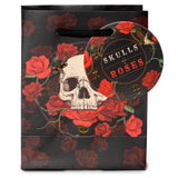 Front view of a small black gift bag, featuring a detailed skull and red roses design with green vines and gold metallic accents. It has black rope handles and a circular 'SKULLS + ROSES' tag attached.