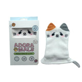 Front view of a white, fluffy cat-shaped makeup remover glove with embroidered features, an orange ear, a grey ear, and a small grey tail, presented upright next to its white retail box with a window showing the glove's face.