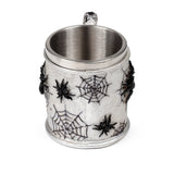 Angled view of a decorative tankard with a textured grey-silver exterior, adorned with black spider web designs and attached black spiders. The metallic interior and textured handle are visible.