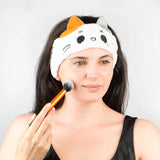 Close-up lifestyle shot of a woman wearing a white plush cat headband with orange and grey ears, applying makeup with a brush.