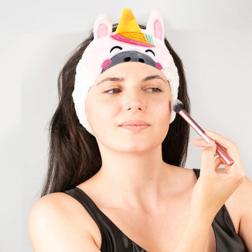 In-context lifestyle shot of a woman wearing a pink plush unicorn headband with a yellow horn and rainbow mane, applying makeup with a brush.
