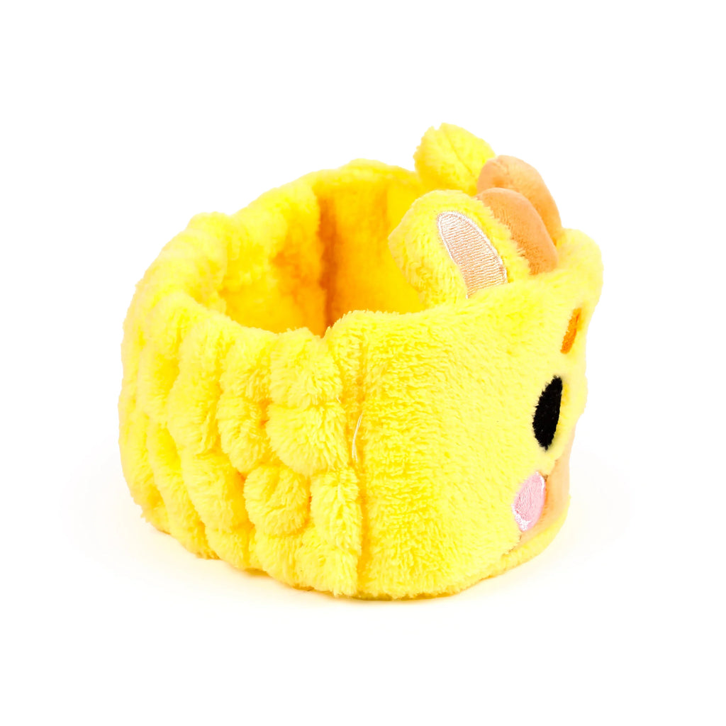 Angled view of a yellow plush beauty headband, featuring an embroidered giraffe face with a black eye, pink cheek, and orange ears.