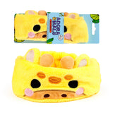Front view of a plush yellow giraffe beauty headband, featuring embroidered brown spots, black eyes, a light brown muzzle, pink cheek details, 3D ears, and horns. It is shown both folded within its light blue cardboard packaging and laid out unfolded on a white background.