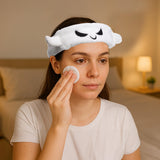 Lifestyle shot of a woman wearing a white plush spooky ghost headband with black embroidered eyes and mouth, applying a cotton pad to her face.