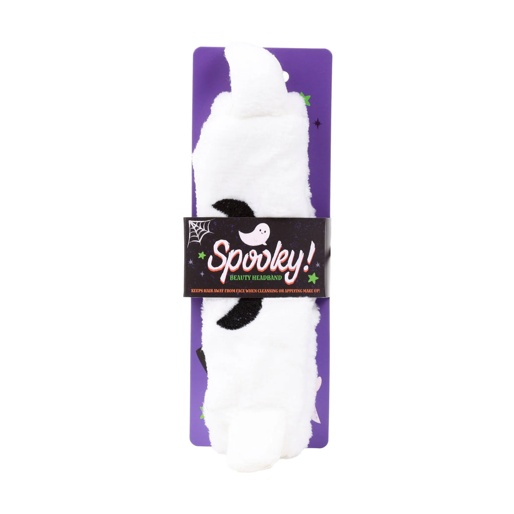 Front view of a white plush ghost-shaped beauty headband with black eyes, packaged on a purple card with a black 'Spooky!' band, spiderweb, and stars.
