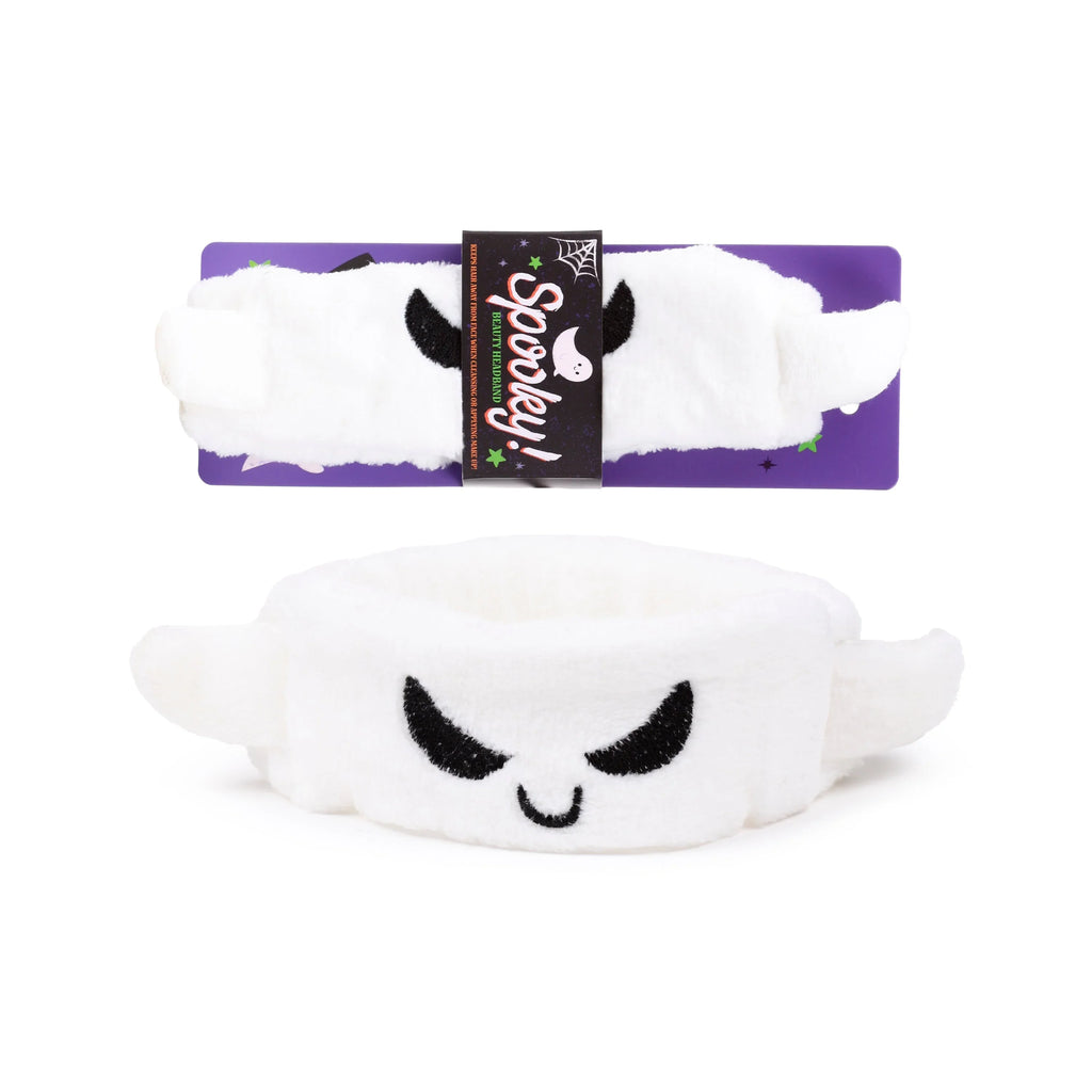 Front view of a white plush ghost beauty headband with black embroidered face, displayed with its purple 'Spooky!' retail packaging.