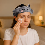 Lifestyle shot of a woman wearing a grey plush bat beauty headband with black ears and an embroidered winking face, applying a product to her cheek with a cotton pad.