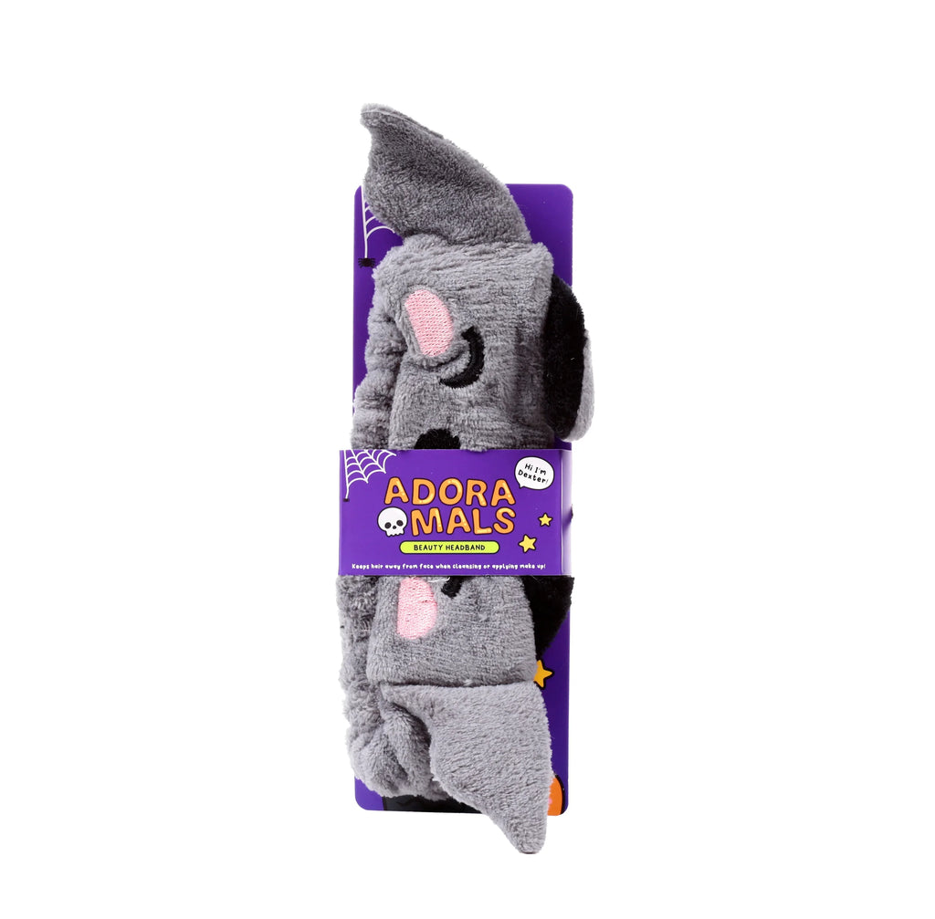 Front view of a grey plush Dexter the Bat beauty headband with pink inner ears and black details, displayed on purple packaging featuring 'Adoramals' text and graphics.