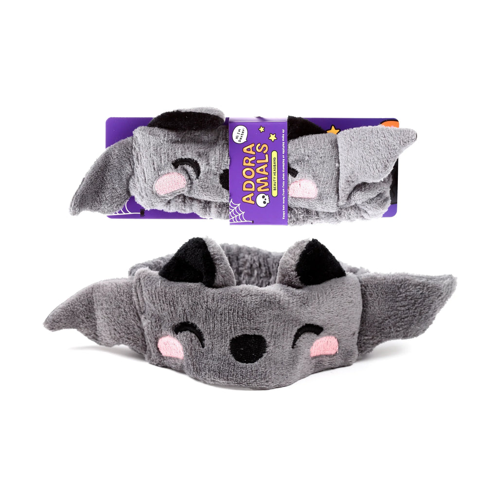 Plush grey Dexter the Bat beauty headband with black ears, embroidered eyes, nose, and pink cheeks. Displayed unpackaged and within purple retail packaging.