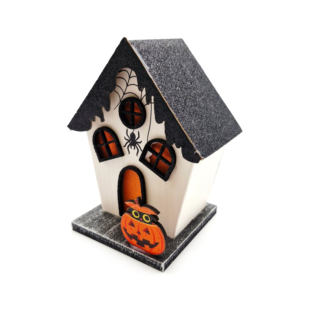 Angled view of a white Halloween decorative house with a glittery black roof and base. It features orange-backed windows and door cutouts, a spiderweb with a spider on one side, and a pumpkin with a black cat at the front.