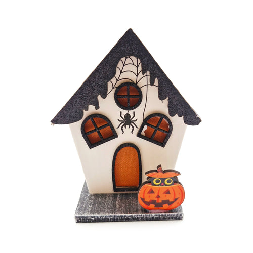 Angled view of a white Halloween spooky house LED decoration, featuring a dark glittery roof, printed spider webs, black spider graphic, and orange-coloured windows and door, with a pumpkin Jack-o'-lantern with a black cat on its base.