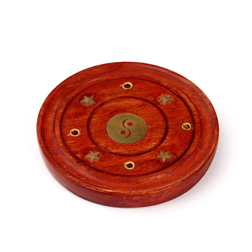 Angled view of a decorative round wooden incense holder ashcatcher with a central brass yin yang design, surrounded by brass stars and incense stick holes.