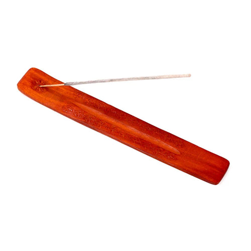 Angled view of a reddish-brown wooden incense holder, showing its intricate carved patterns, central ash-catching groove, and an unlit incense stick inserted.