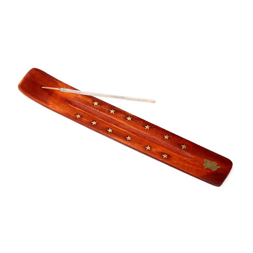 Angled overhead view of a reddish-brown mango wood incense holder with golden star inlays along the ash-catching groove and a decorative golden crown emblem at one end. An unlit grey incense stick is inserted.