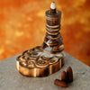 Angled view of a lit mango wood backflow incense burner, showing white smoke cascading over its tiered dark wooden design and into the intricate carved patterns on its base. Three brown incense cones rest nearby on a grey surface.