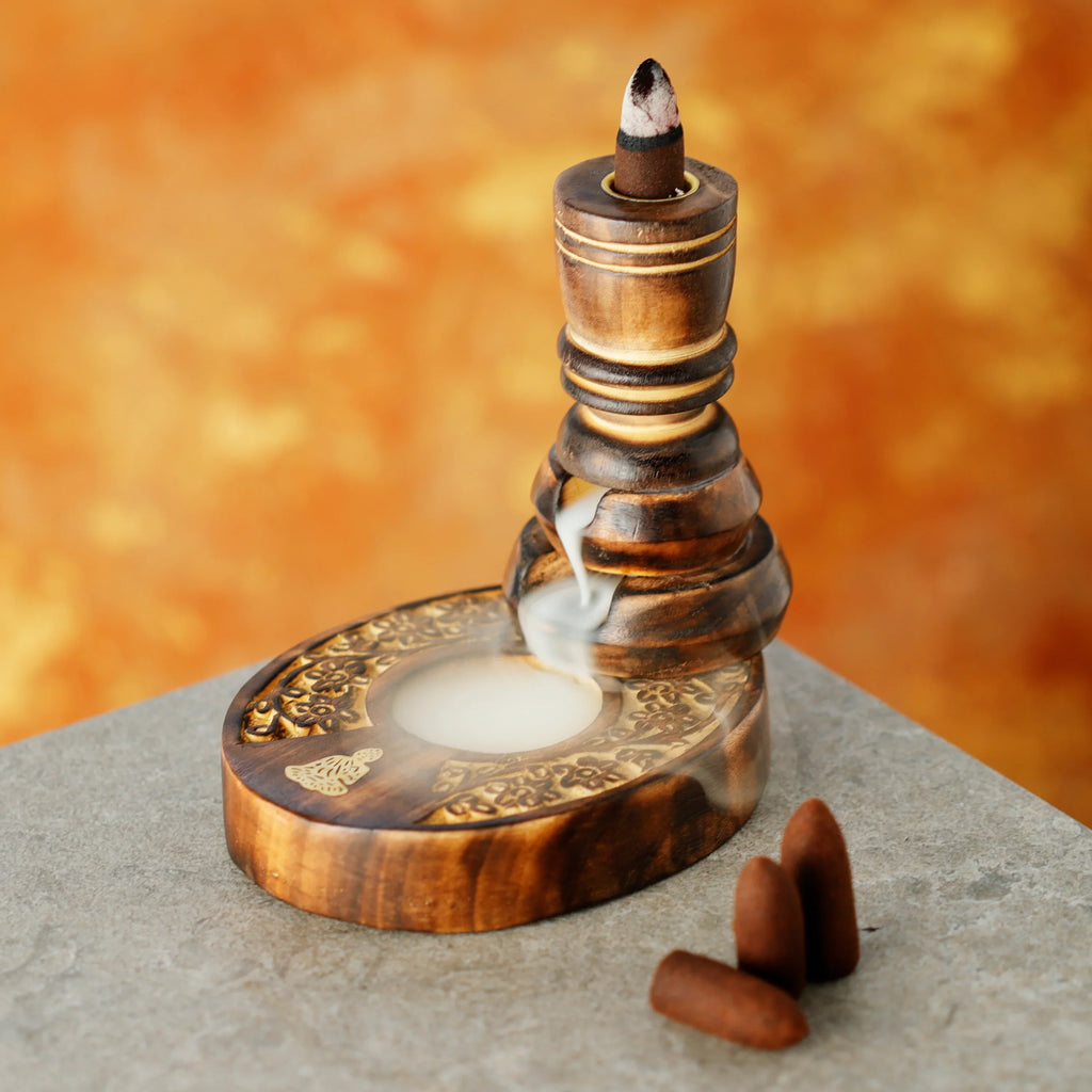 Angled view of a dark carved wooden backflow incense burner, with smoke flowing down its tiered column into a base featuring intricate floral patterns. A burning incense cone is visible at the top, and three unlit cones lie on the grey surface next to it.