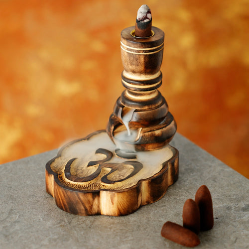 Angled view of a dark mango wood backflow incense burner, with a lit cone releasing smoke that flows over its Om-carved base. Three unlit incense cones sit beside it.