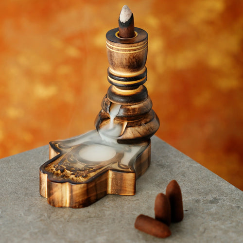 Angled view of a carved mango wood backflow incense burner with an intricately detailed Hamsa hand-shaped base, showing smoke cascading from the lit cone into the base. Three unlit brown backflow incense cones are visible on the grey surface.