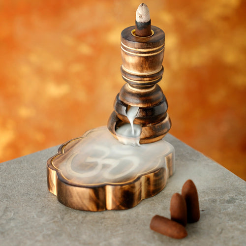Angled view of a carved Om mango wood backflow incense burner, with white smoke cascading over the Om symbol on its base. Three brown incense cones are placed next to it on a grey surface.