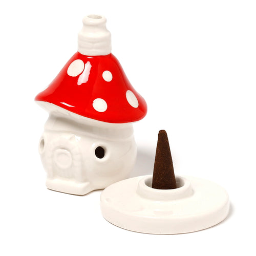 Angled view of a white ceramic fairy toadstool house incense burner with a red spotted cap, displayed next to its matching white ceramic base holding a dark incense cone.