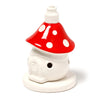 A ceramic incense cone burner shaped like a fairy toadstool house, featuring a glossy red cap with white spots and a central opening for incense, a cream-coloured body with a small circular vent, and a flat circular base.