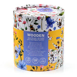 Angled view of a cylindrical wooden jigsaw puzzle packaging, with a vibrant floral and bee meadow design. The prominent purple label reads 'WOODEN JIGSAW' and displays icons for '130 pieces' and 'wooden puzzle'.