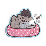 Partially assembled 130-piece wooden jigsaw puzzle, depicting Pusheen the Cat and two kittens in a pink spotted bath with a light blue border, with loose pieces scattered, overhead view.