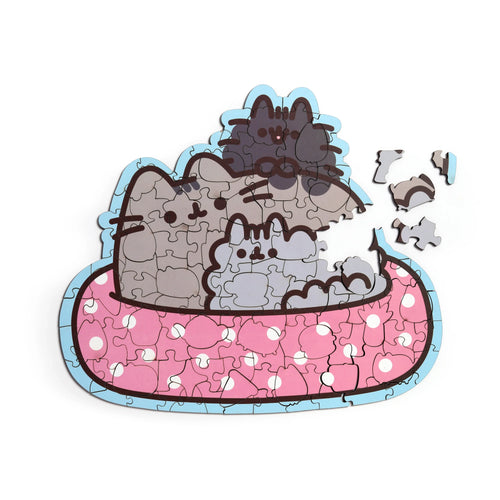 Partially assembled 130-piece wooden jigsaw puzzle, depicting Pusheen the Cat and two kittens in a pink spotted bath with a light blue border, with loose pieces scattered, overhead view.