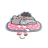 Angled overhead view of an incomplete wooden jigsaw puzzle depicting grey Pusheen the Cat characters on a pink polka-dot cushion, with three loose wooden pieces lying beneath.